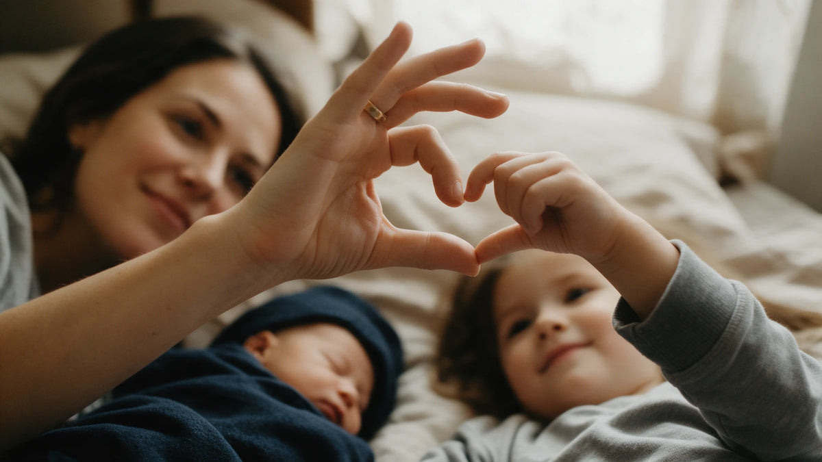 A mother lies on a bed with her newborn and toddler, she and the older child touching fingertips to form a heart.